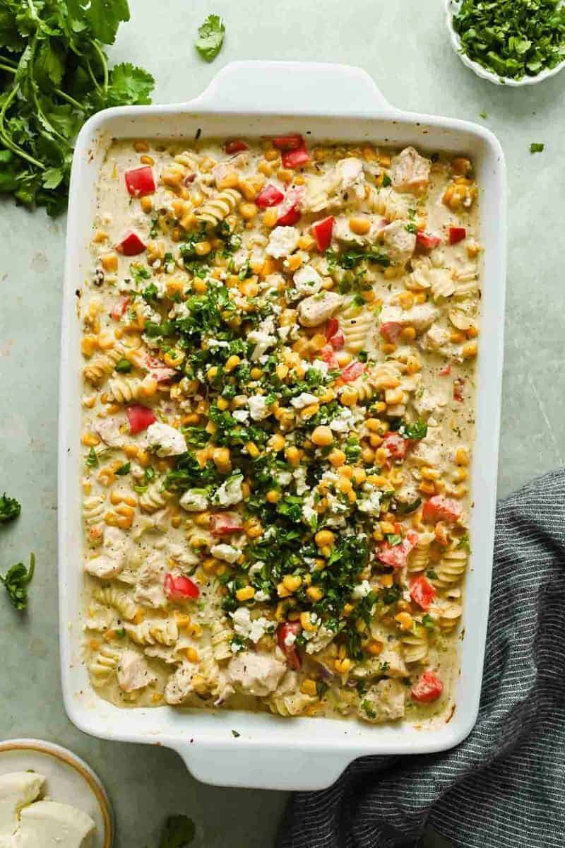 A white baking dish filled with a creamy pasta bake with rotini, corn, diced tomatoes, herbs, and cheese, topped with chopped cilantro and crumbled cheese.