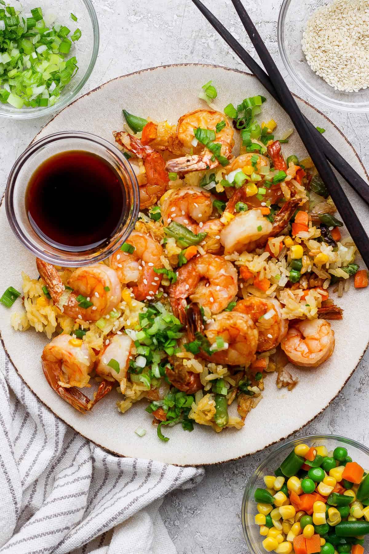 A plate of shrimp fried rice with mixed vegetables, topped with chopped green onions, served with soy sauce and chopsticks. Small bowls of sesame seeds and vegetables are beside the plate.