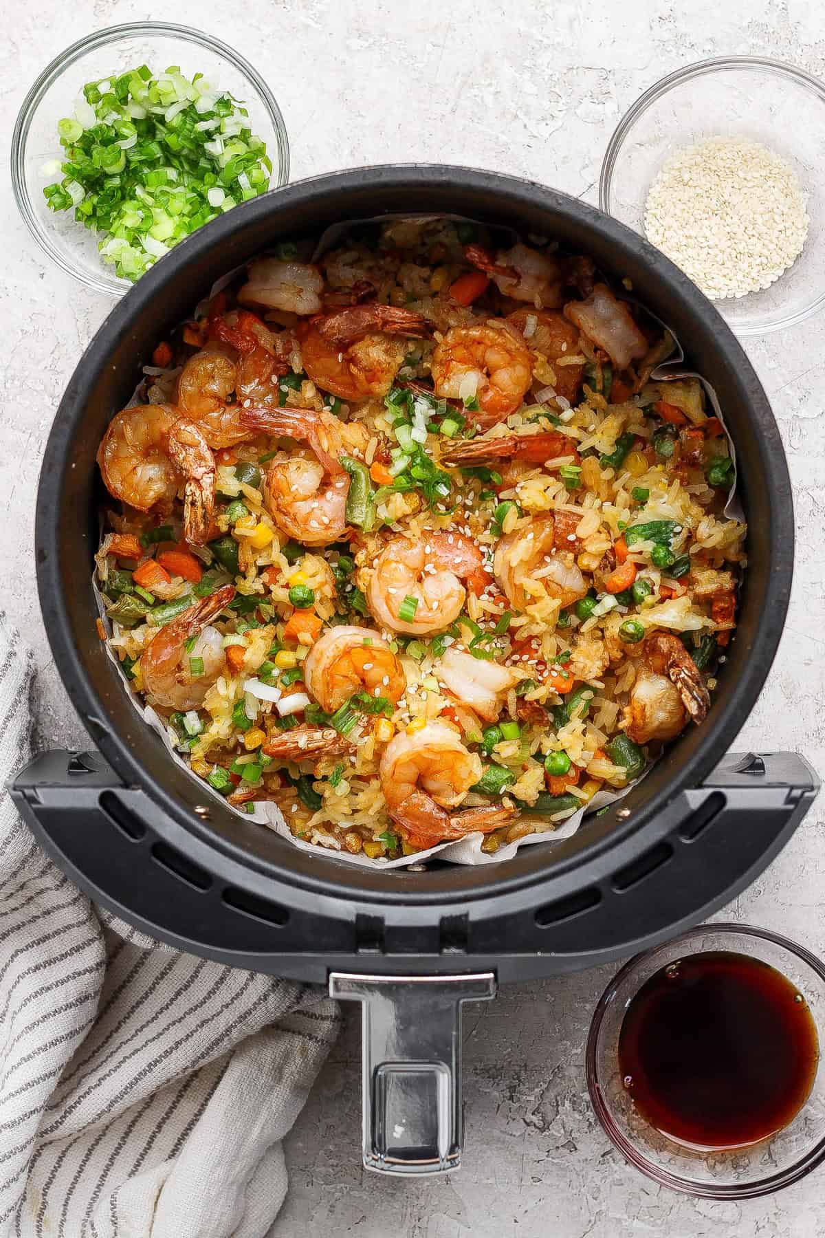 Cooked shrimp fried rice in an air fryer basket, garnished with chopped green onions, with bowls of sesame seeds, green onions, and a dark sauce nearby.