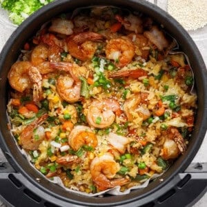 A close-up of a cooked shrimp fried rice dish with mixed vegetables, served in a black bowl and garnished with chopped green onions and sesame seeds.