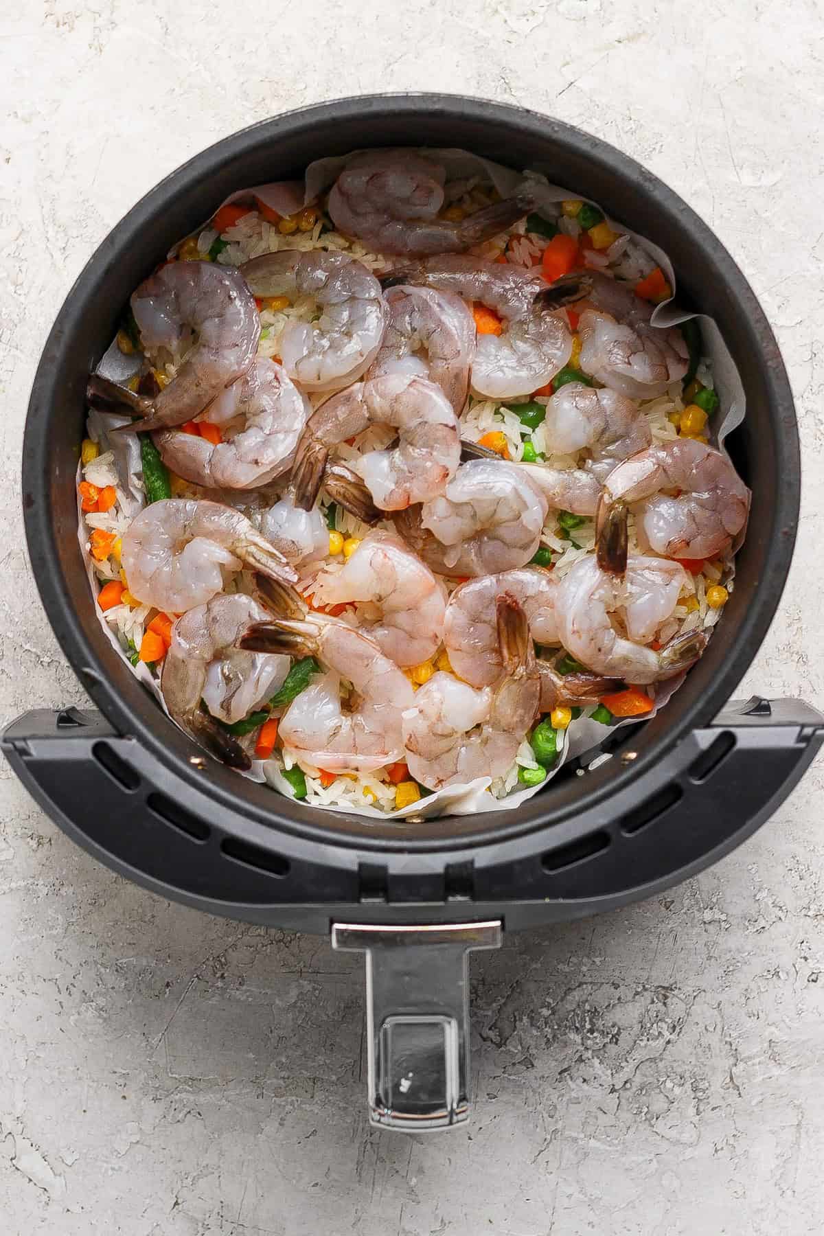 Raw shrimp arranged on top of mixed vegetables and rice in the basket of an air fryer.