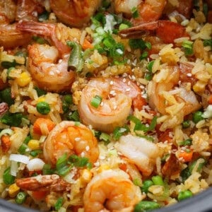 Close-up of shrimp fried rice with peas, corn, green onions, and sesame seeds, served in a black bowl or container.