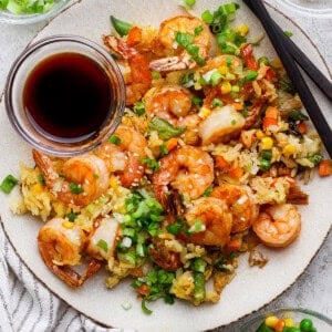 A plate of shrimp fried rice with mixed vegetables, topped with chopped green onions, served with soy sauce and chopsticks. Small bowls of sesame seeds and vegetables are beside the plate.