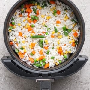 An air fryer basket filled with uncooked white rice and mixed vegetables including carrots, peas, corn, and green beans.