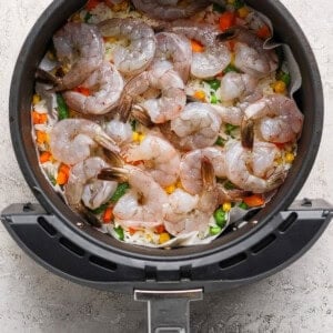 Raw shrimp arranged on top of mixed vegetables and rice in the basket of an air fryer.