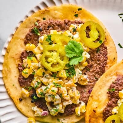 A close-up of a taco with ground beef, creamy corn salad, pickled jalapeño slices, and a sprig of cilantro on a white plate.