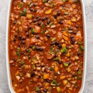 A rectangular white casserole dish filled with a chunky mixture of beans, tomatoes, rice, and vegetables in a red sauce on a textured surface.
