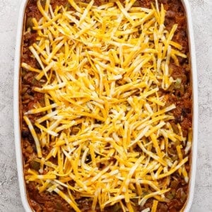 A rectangular baking dish filled with a casserole topped with a layer of shredded yellow and white cheese, set on a light textured surface.