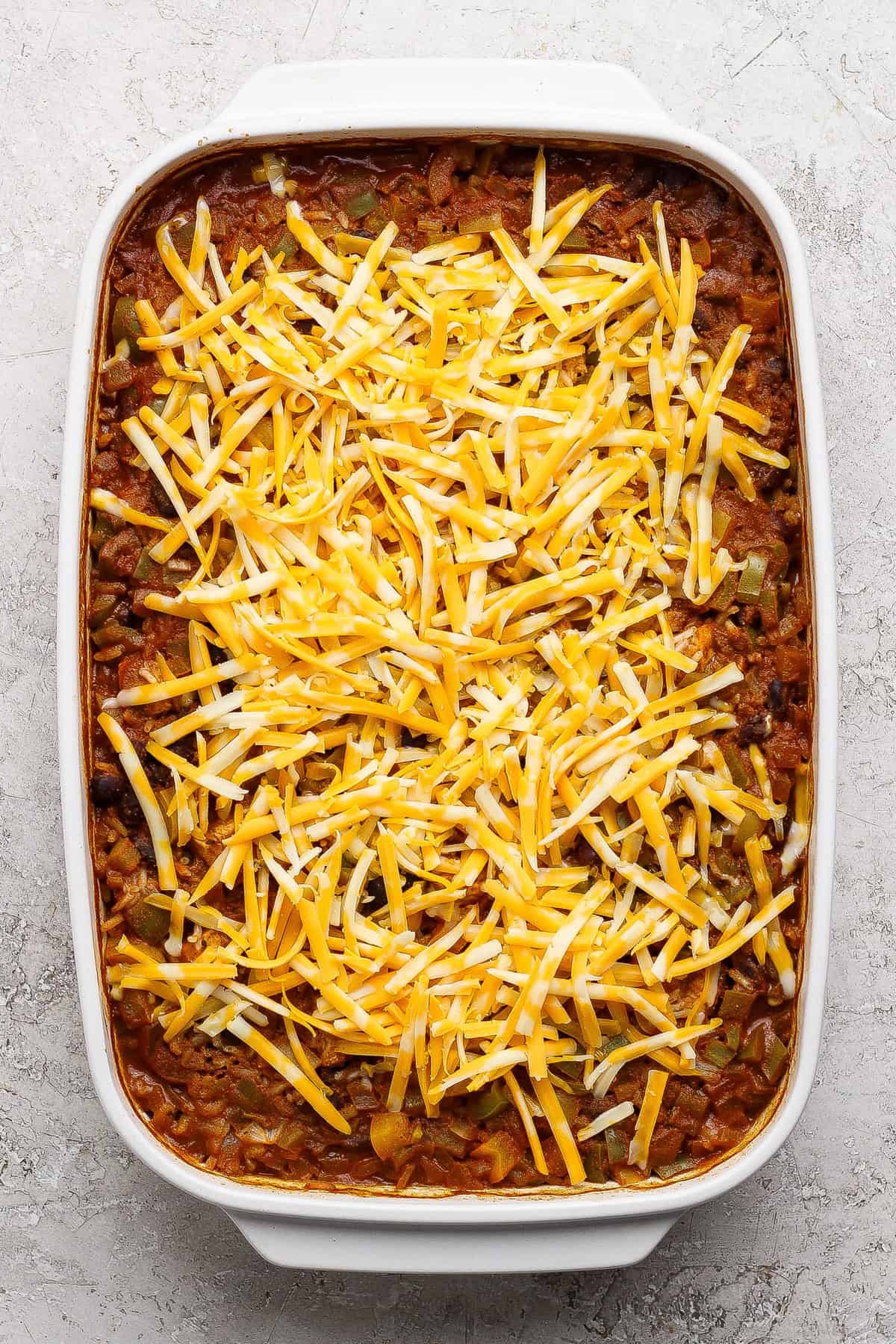 A rectangular baking dish filled with a casserole topped with a layer of shredded yellow and white cheese, set on a light textured surface.