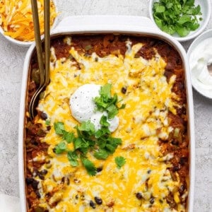 A casserole dish filled with a baked cheesy Mexican dish, topped with sour cream and fresh cilantro, surrounded by bowls of shredded cheese, cilantro, and sour cream.