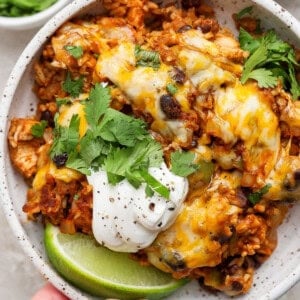 A hand holds a bowl of cheesy chicken and rice topped with sour cream, fresh cilantro, and a lime wedge; small bowls of cilantro and lime are in the background.