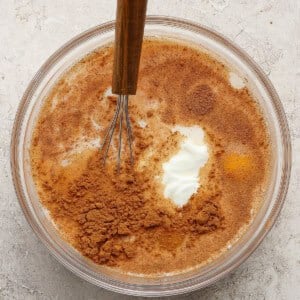 A glass bowl with a whisk contains milk, eggs, a dollop of yogurt, and a layer of cinnamon on top, sitting on a light textured surface.