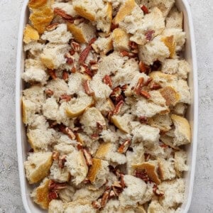 A white baking dish filled with chunks of bread and chopped pecans on a light-colored surface.
