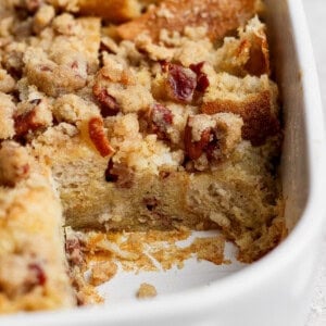 A close-up of a baked bread pudding with a crumbly topping in a white dish, with a corner piece missing.