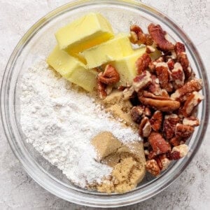 A glass bowl containing cubed butter, chopped pecans, flour, and brown sugar on a light textured surface.