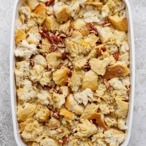 A white baking dish filled with unbaked bread pudding made of torn bread pieces and pecans on a light textured surface.