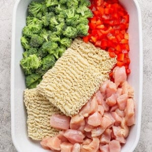 A white baking dish containing raw broccoli florets, diced red bell pepper, two blocks of dry ramen noodles, and chunks of raw chicken, all arranged separately.