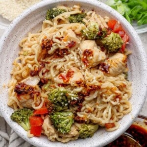 Bowl of ramen noodles with pieces of chicken, broccoli, red bell peppers, sesame seeds, and a chili oil sauce.