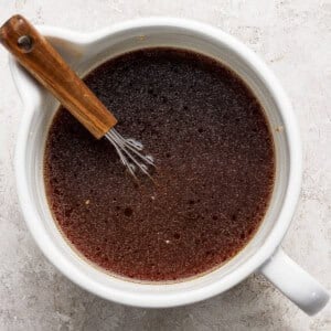 A small metal whisk rests in a white ceramic bowl filled with a dark brown liquid mixture on a light textured surface.