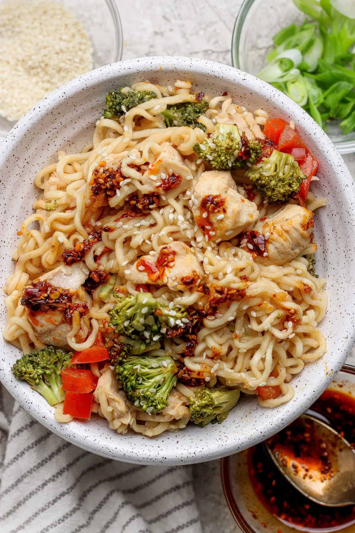 A bowl of ramen noodles mixed with chicken, broccoli, and red bell peppers, topped with chili oil and sesame seeds, with green onions and sauces on the side.