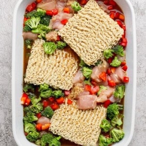 Uncooked ramen noodles, raw chicken pieces, chopped broccoli, and diced red bell peppers in a white rectangular baking dish.
