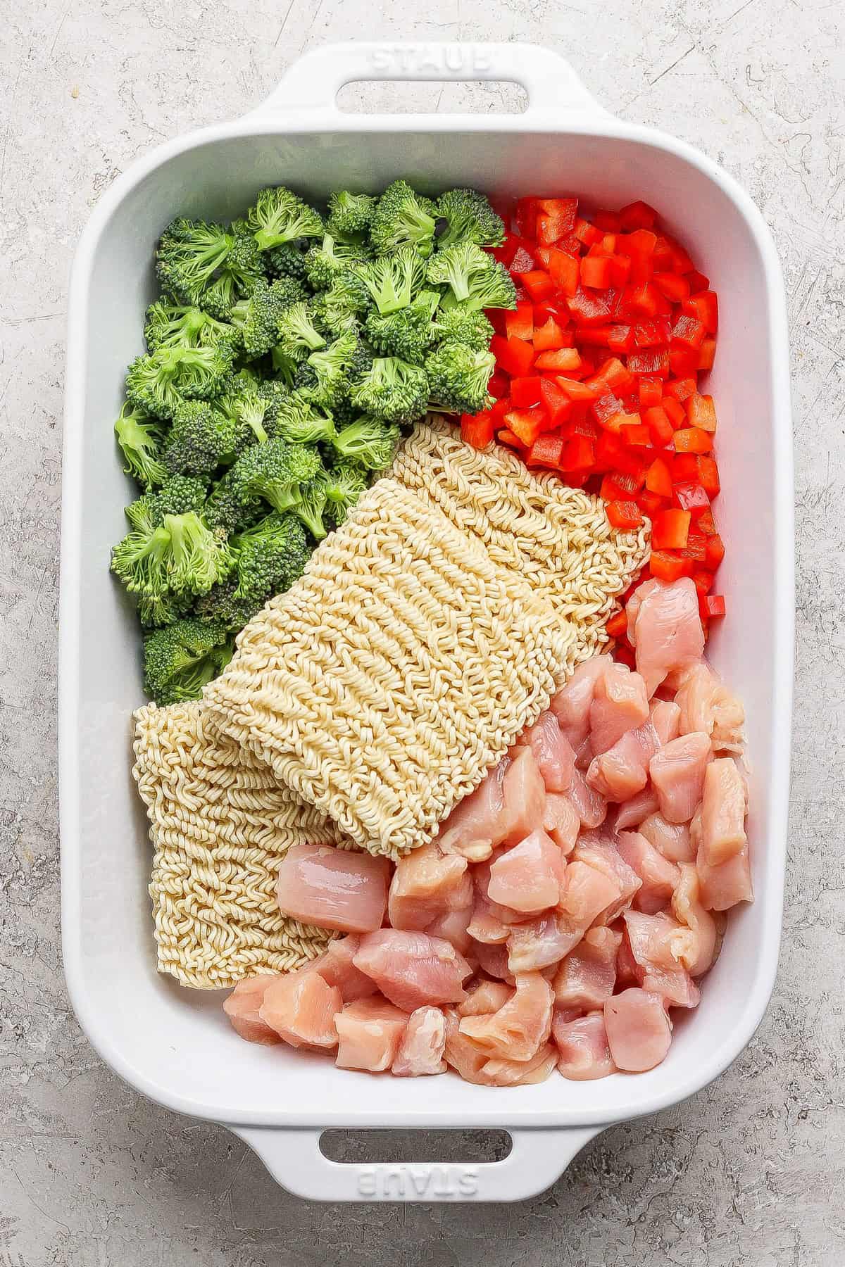 A white baking dish containing raw broccoli florets, diced red bell pepper, two blocks of dry ramen noodles, and chunks of raw chicken, all arranged separately.