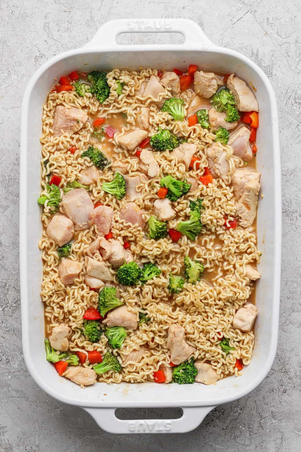 A white baking dish filled with cooked ramen noodles, pieces of chicken, broccoli florets, and red bell pepper in a light broth.