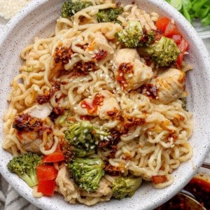 A bowl of ramen noodles mixed with chicken, broccoli, and red bell peppers, topped with chili oil and sesame seeds, with green onions and sauces on the side.