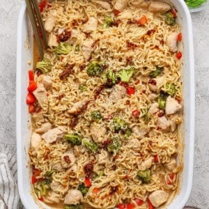 Baked ramen casserole with chicken, broccoli, red bell peppers, and sesame seeds in a white baking dish, with a gold serving spoon and garnishes on the side.