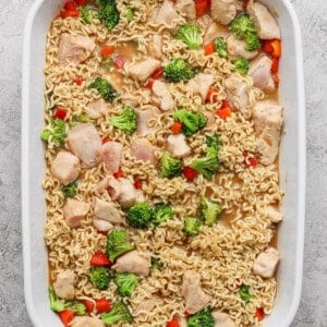 A white baking dish filled with cooked ramen noodles, pieces of chicken, broccoli florets, and red bell pepper in a light broth.
