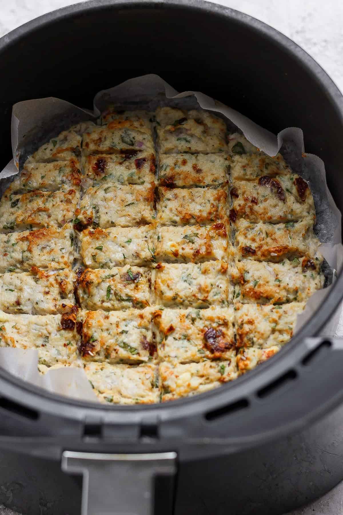 A round dish of baked, sliced savory bars with herbs and browned spots, lined with parchment paper, inside an air fryer basket.