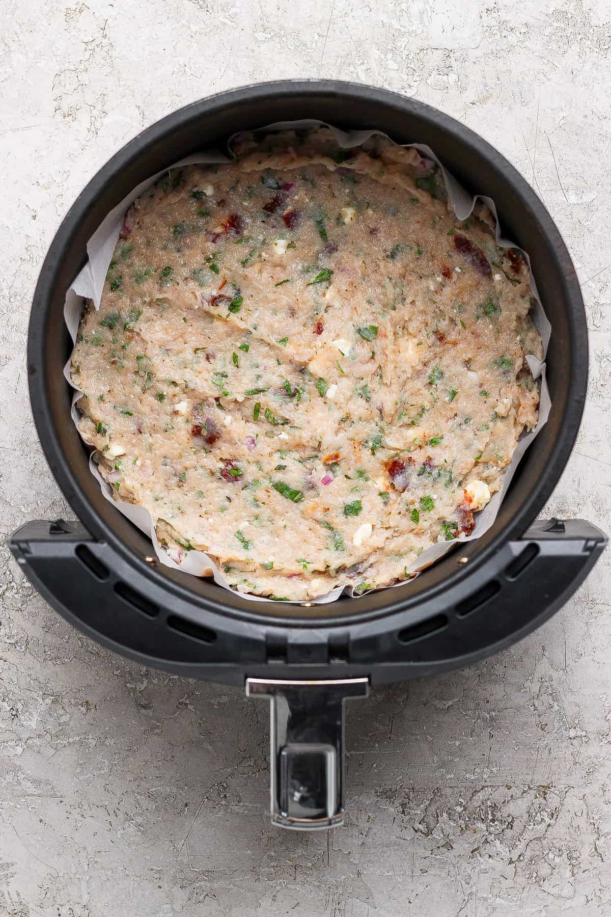 Uncooked mixture of herbs, vegetables, and cheese pressed into a round baking dish lined with parchment paper inside an air fryer basket.