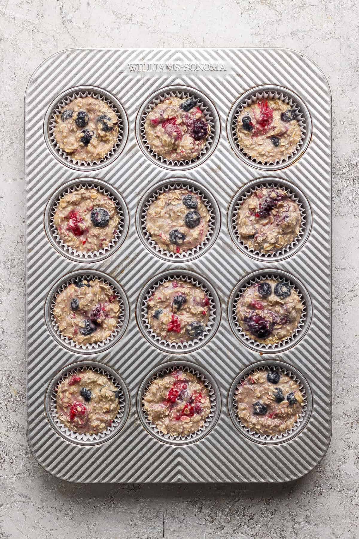 A muffin tin with twelve paper liners filled with unbaked oatmeal batter and mixed berries, placed on a light textured surface.