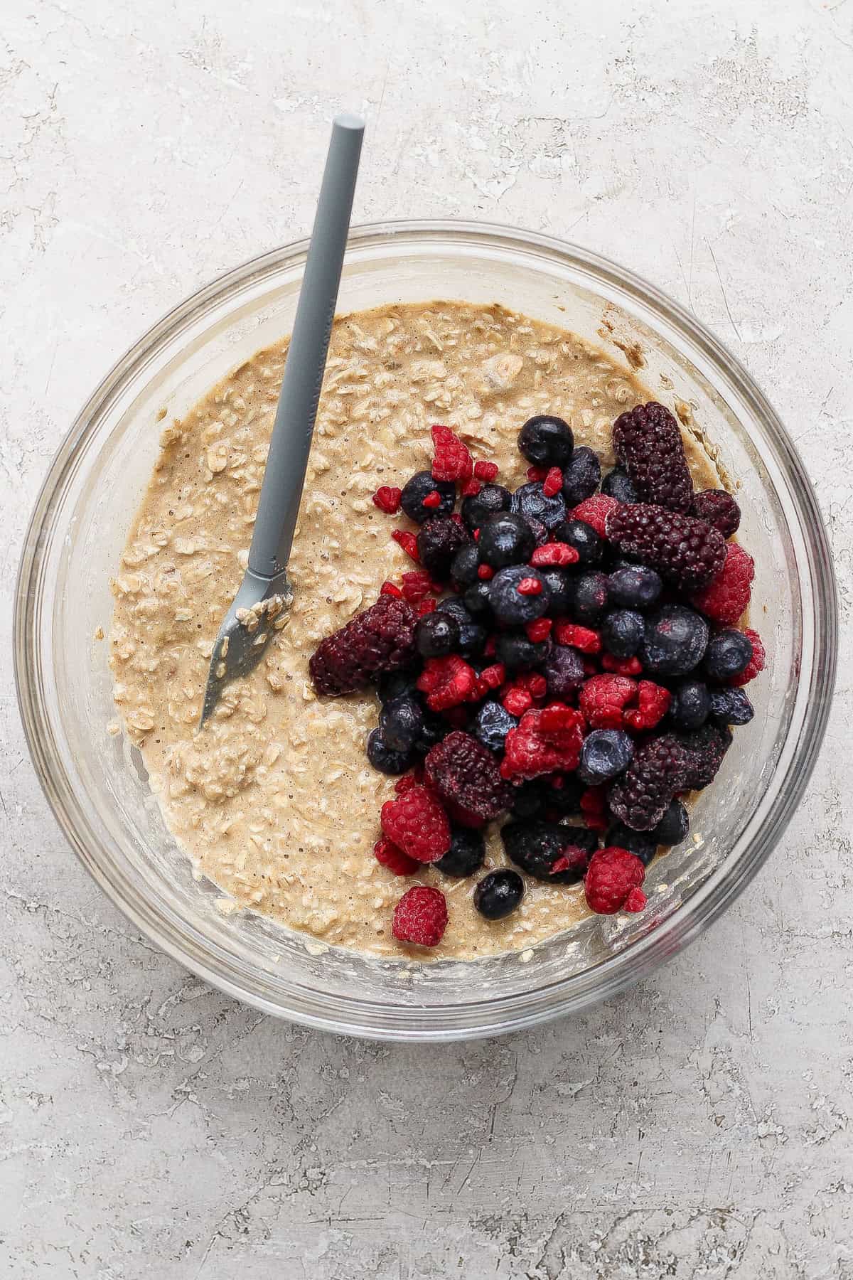 A glass bowl containing oatmeal batter with a pile of mixed berries and a spoon resting inside, placed on a light textured surface.