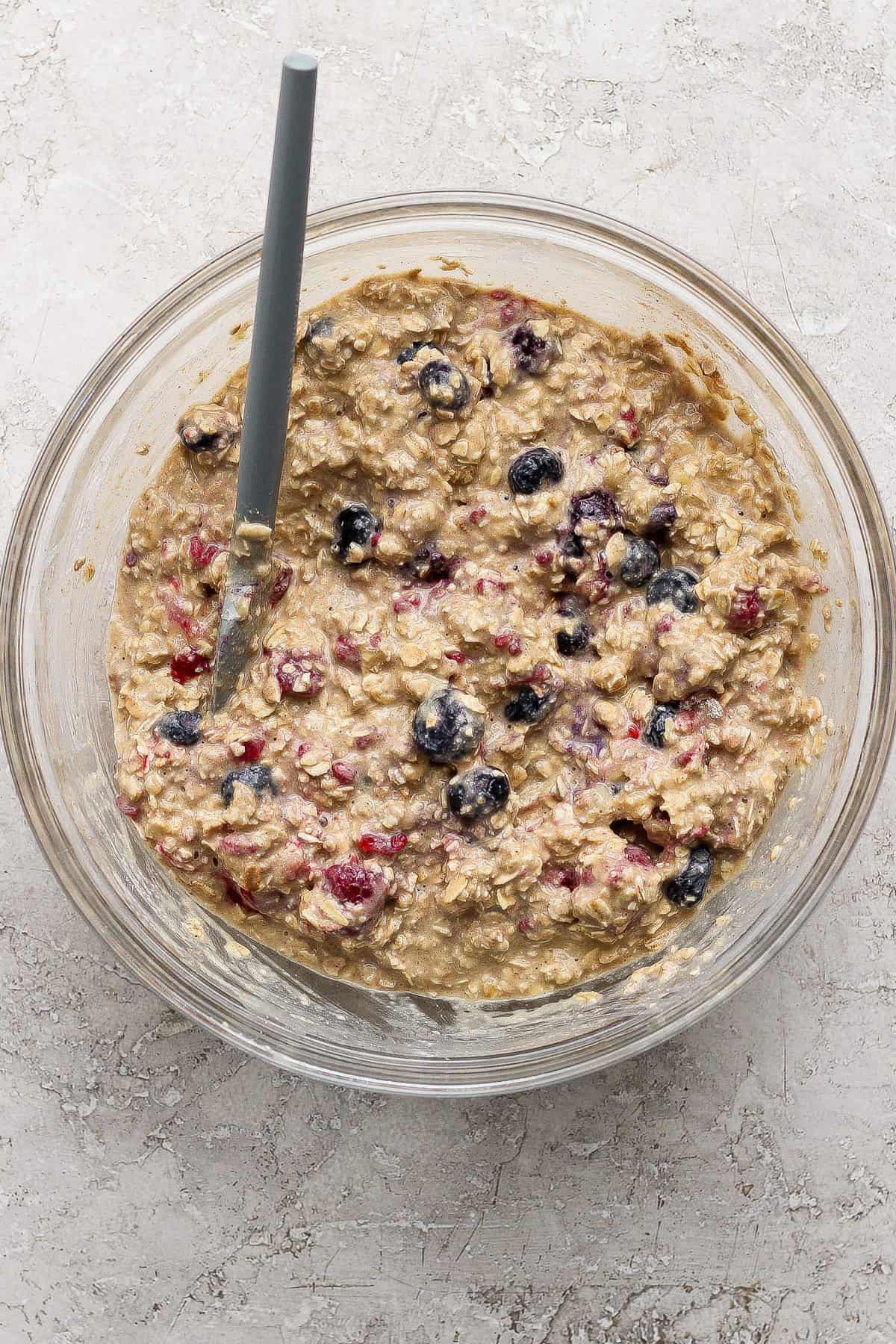 A glass bowl filled with oatmeal batter mixed with berries, with a spoon resting inside, on a light textured surface.