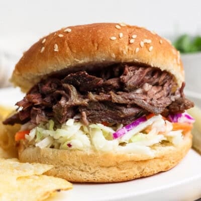 Shredded beef sandwich with coleslaw served on a sesame seed bun, with potato chips on the side.