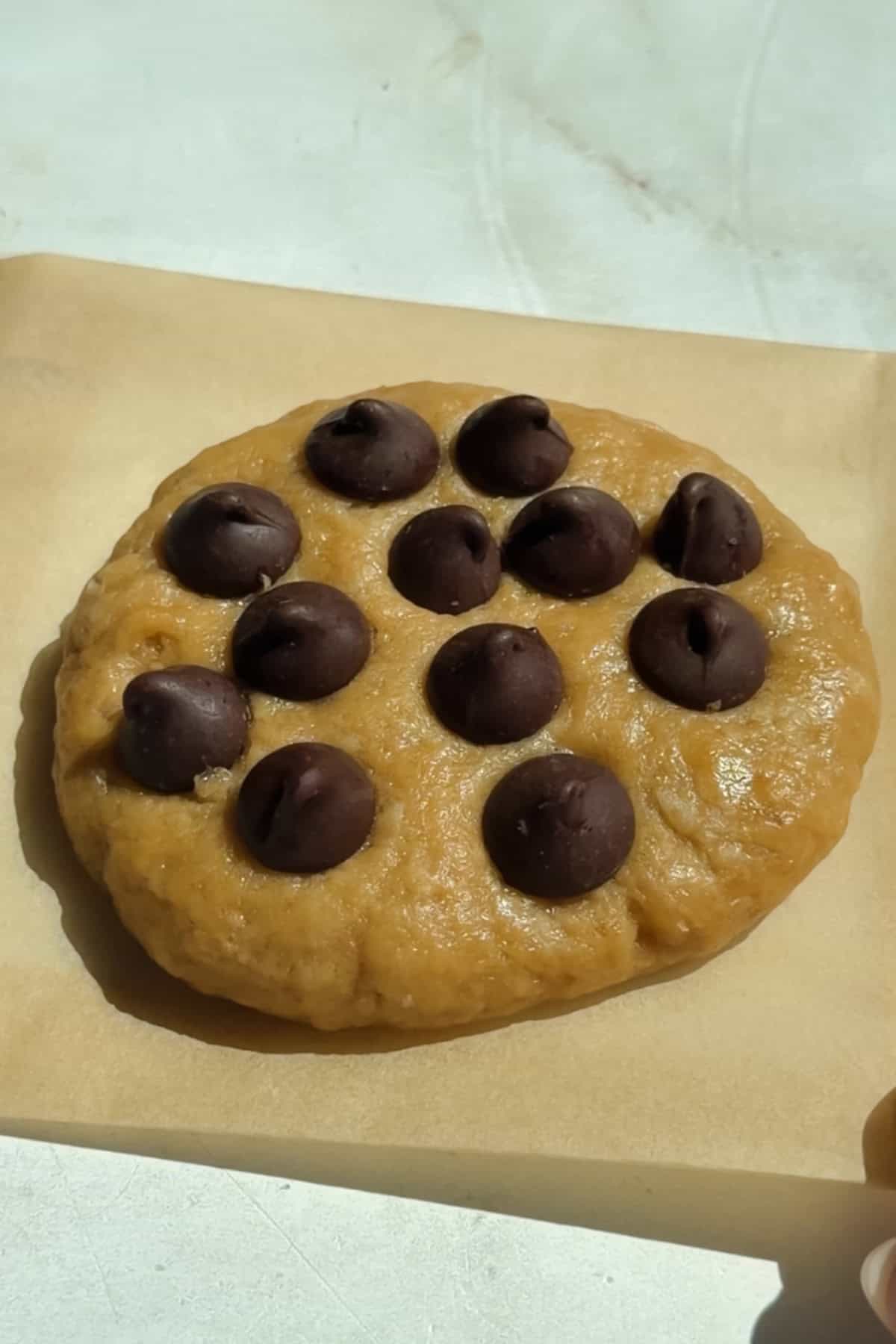 A round piece of cookie dough topped with chocolate chips sits on a sheet of parchment paper.