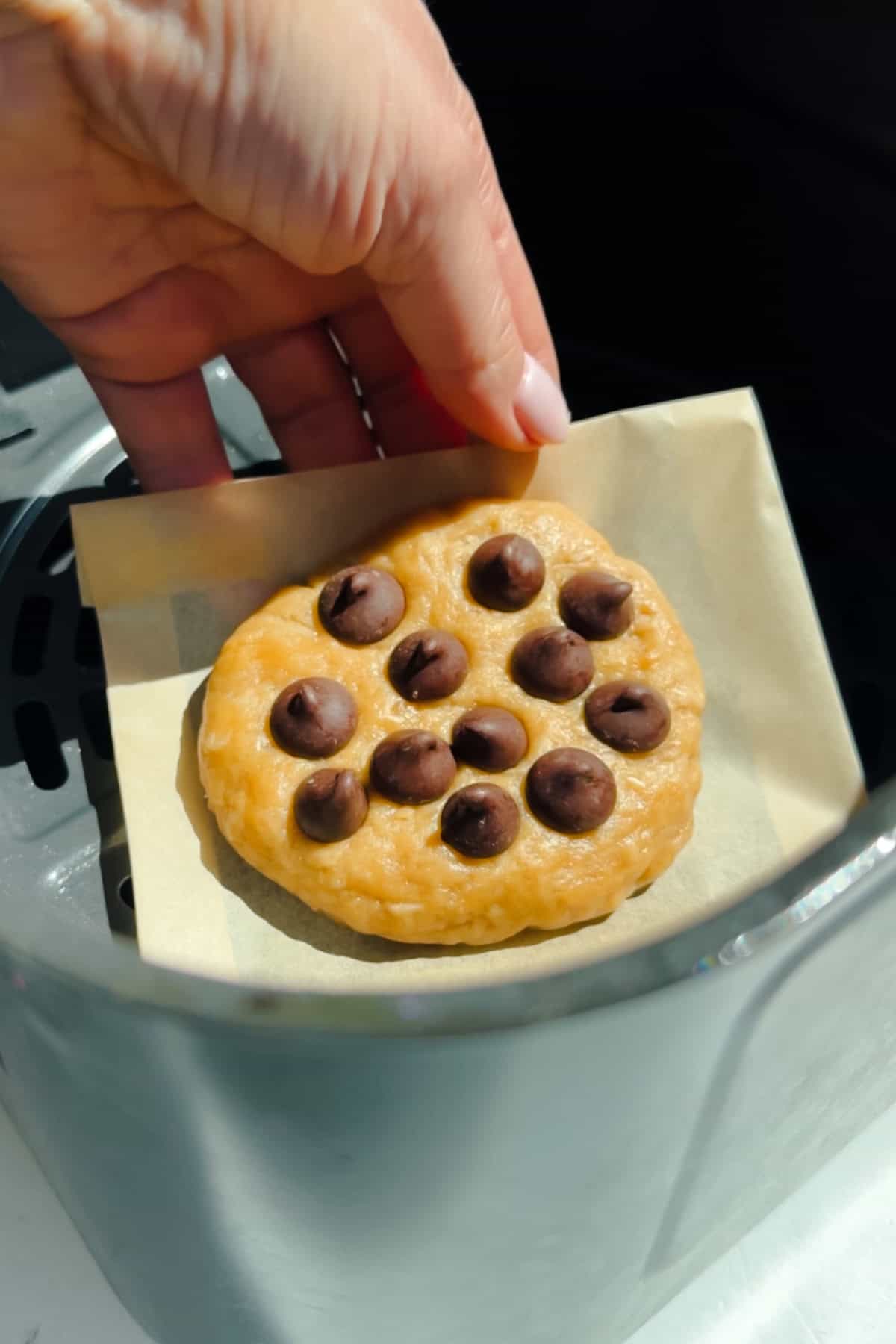 A hand is placing a round chocolate chip cookie dough on a piece of parchment paper inside an air fryer basket.