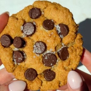 A person holding a chocolate chip cookie topped with coarse sea salt; the cookie has a golden-brown color and visible chocolate chips.