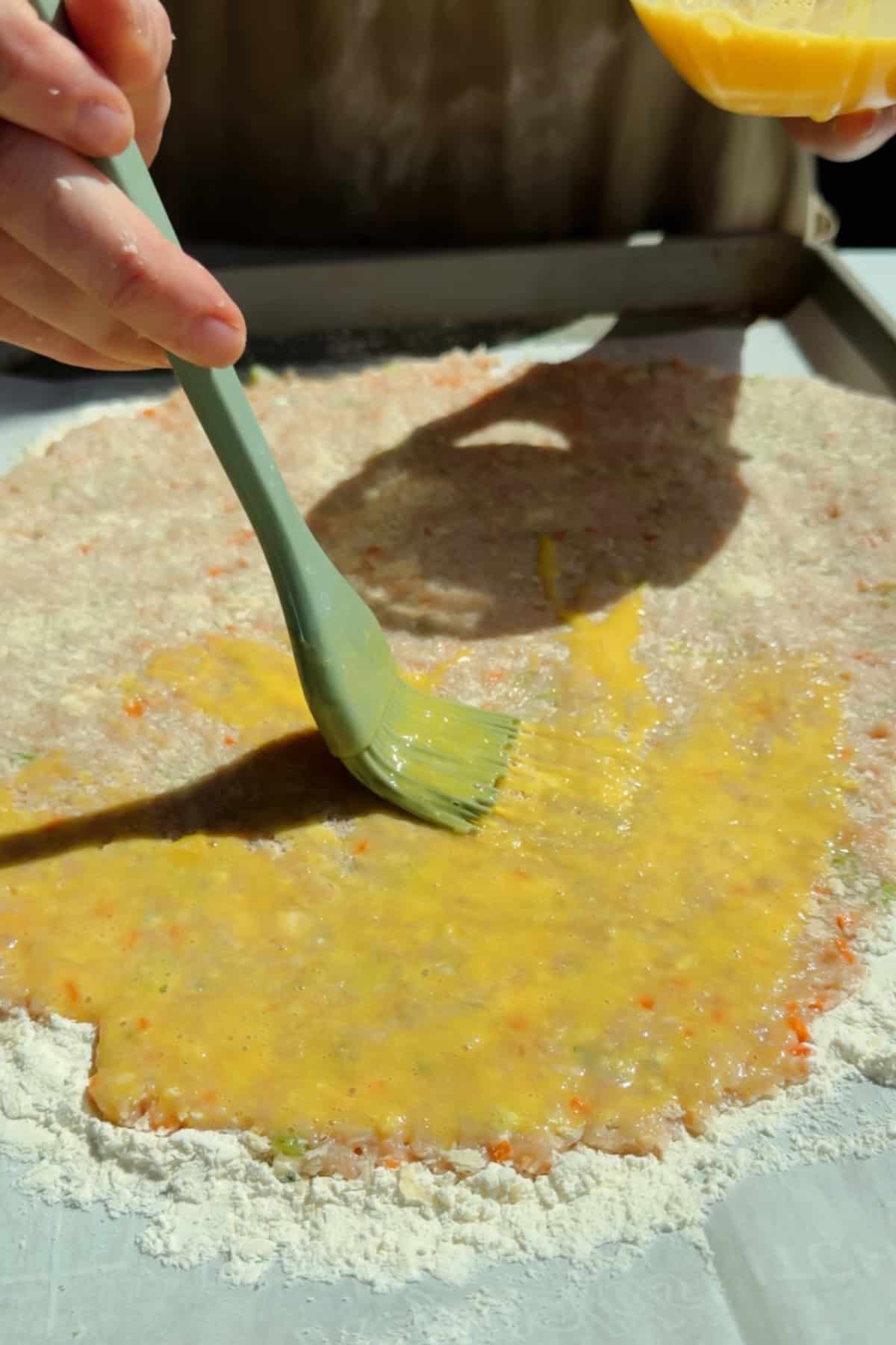 Brushing egg wash over chicken protein bar mixture on a floured parchment-lined surface