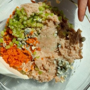 Mixing ground chicken, carrots, celery, blue cheese crumbles, and seasonings in a glass bowl for protein bars