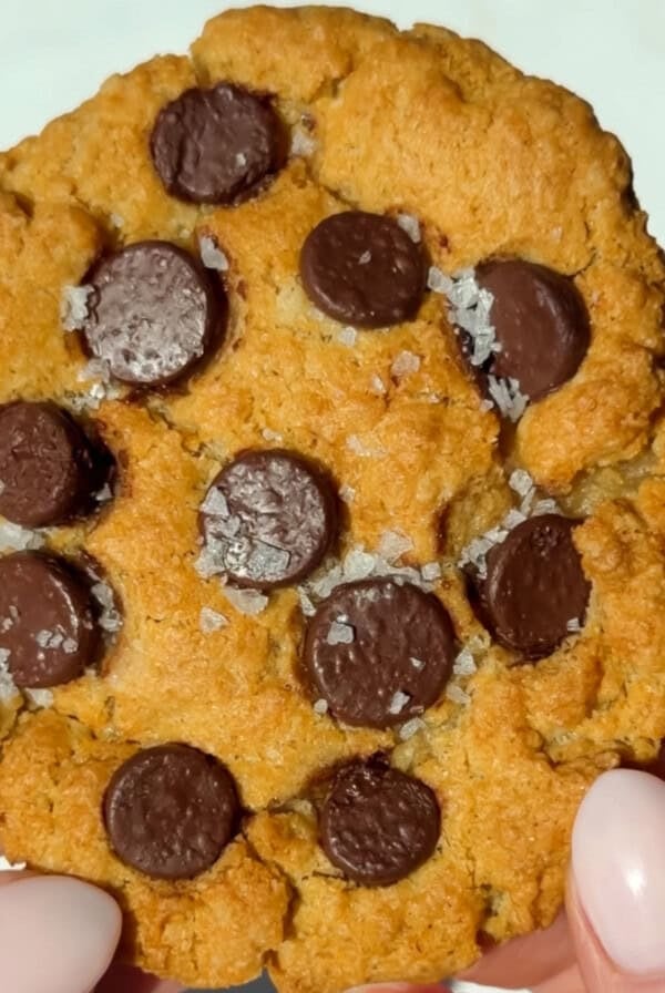 A person holding a chocolate chip cookie topped with coarse sea salt; the cookie has a golden-brown color and visible chocolate chips.
