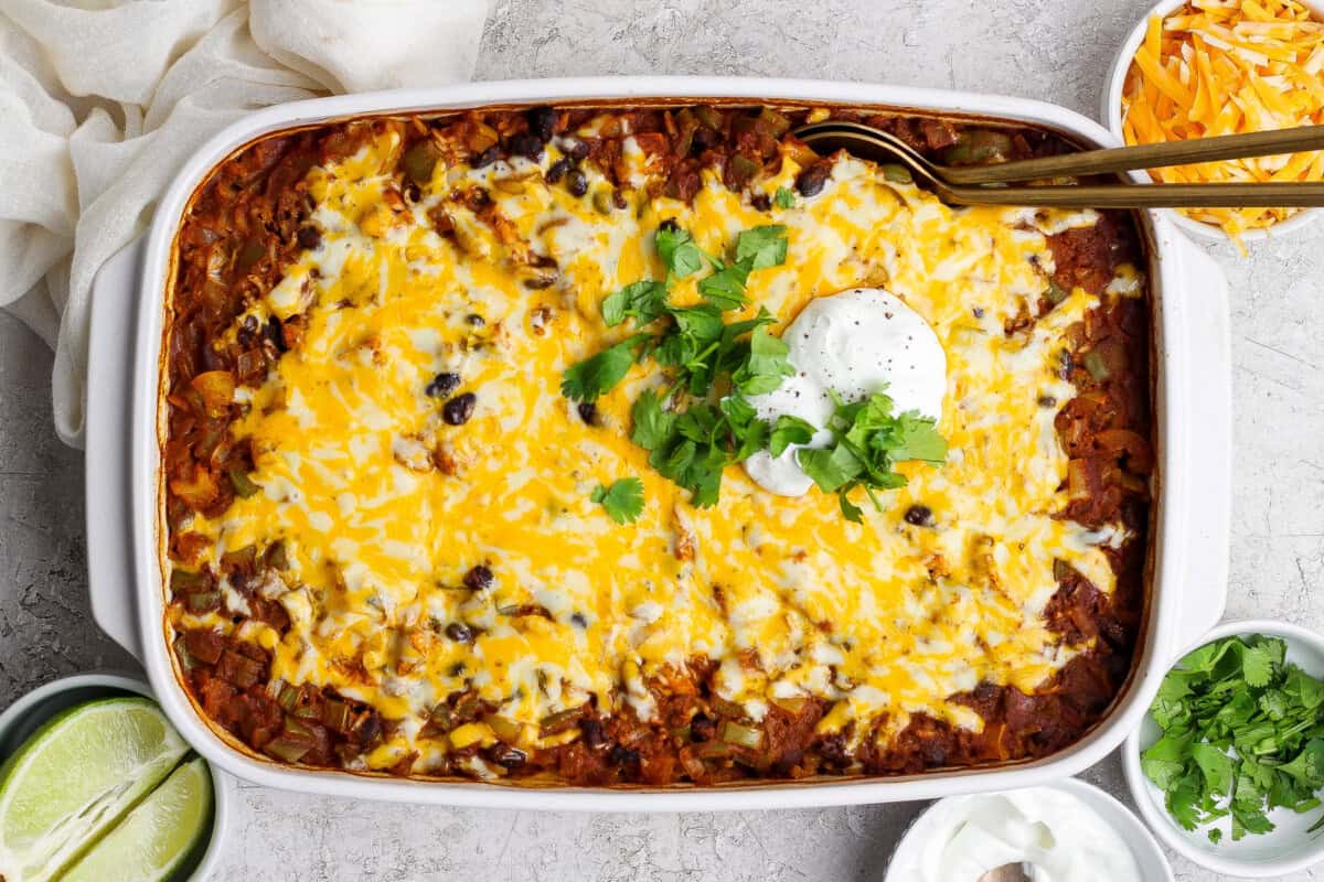 A baked casserole topped with melted cheese, black beans, sour cream, and chopped cilantro, surrounded by bowls of lime, shredded cheese, and extra sour cream.