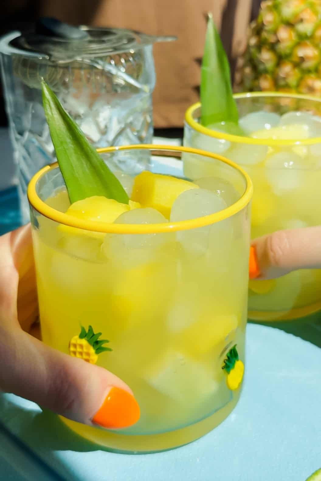 Two hands holding glasses filled with ice, yellow drink, pineapple chunks, and a pineapple leaf garnish, decorated with small pineapple designs.