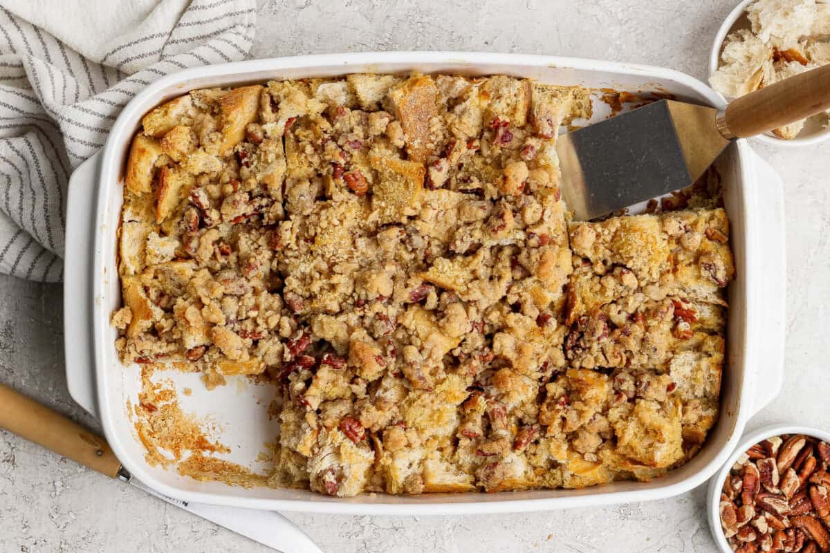 Baked bread pudding with a crumbly topping in a white rectangular dish, partially served with a spatula, surrounded by bowls of pecans and coconut.
