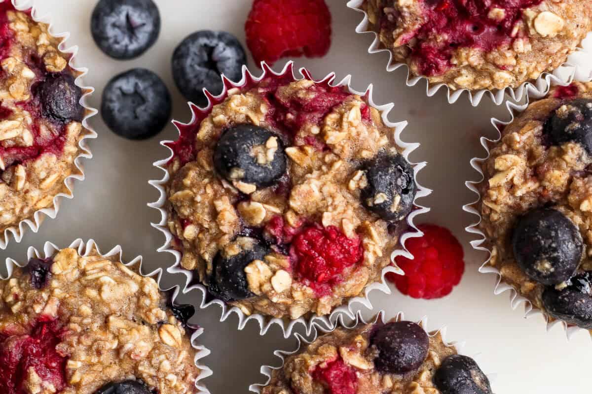 Get your meal prep on and make these easy Triple Berry Baked Oatmeal Cups for the week's breakfast. This baked oatmeal recipe is made with 3 kinds of berries, rolled oats, and ground oat flour making this a better-for-you breakfast or snack! Baked oatmeal muffins topped with blueberries and raspberries, arranged on a white surface with additional berries nearby.