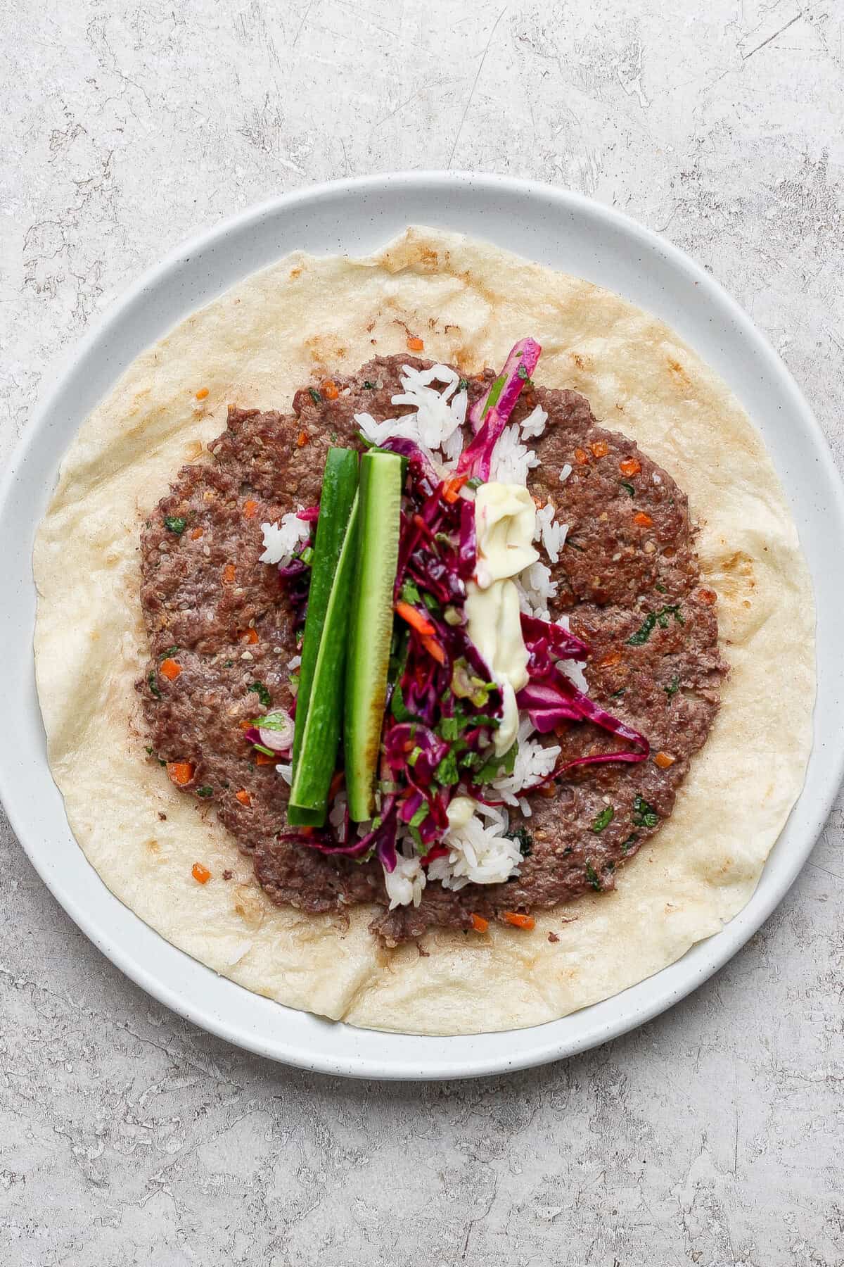 A plate with a flatbread topped with ground meat, rice, red cabbage slaw, cucumber sticks, and a drizzle of white sauce.