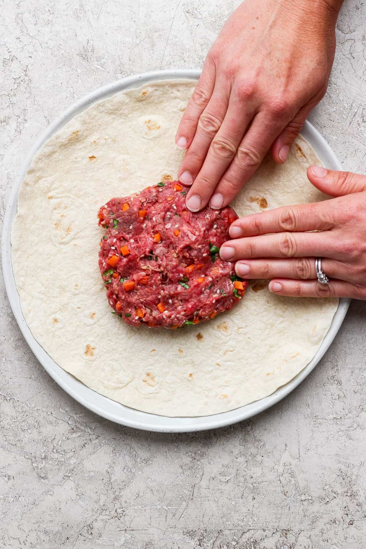 Hands pressing raw ground meat mixture with chopped vegetables onto a large tortilla on a plate.