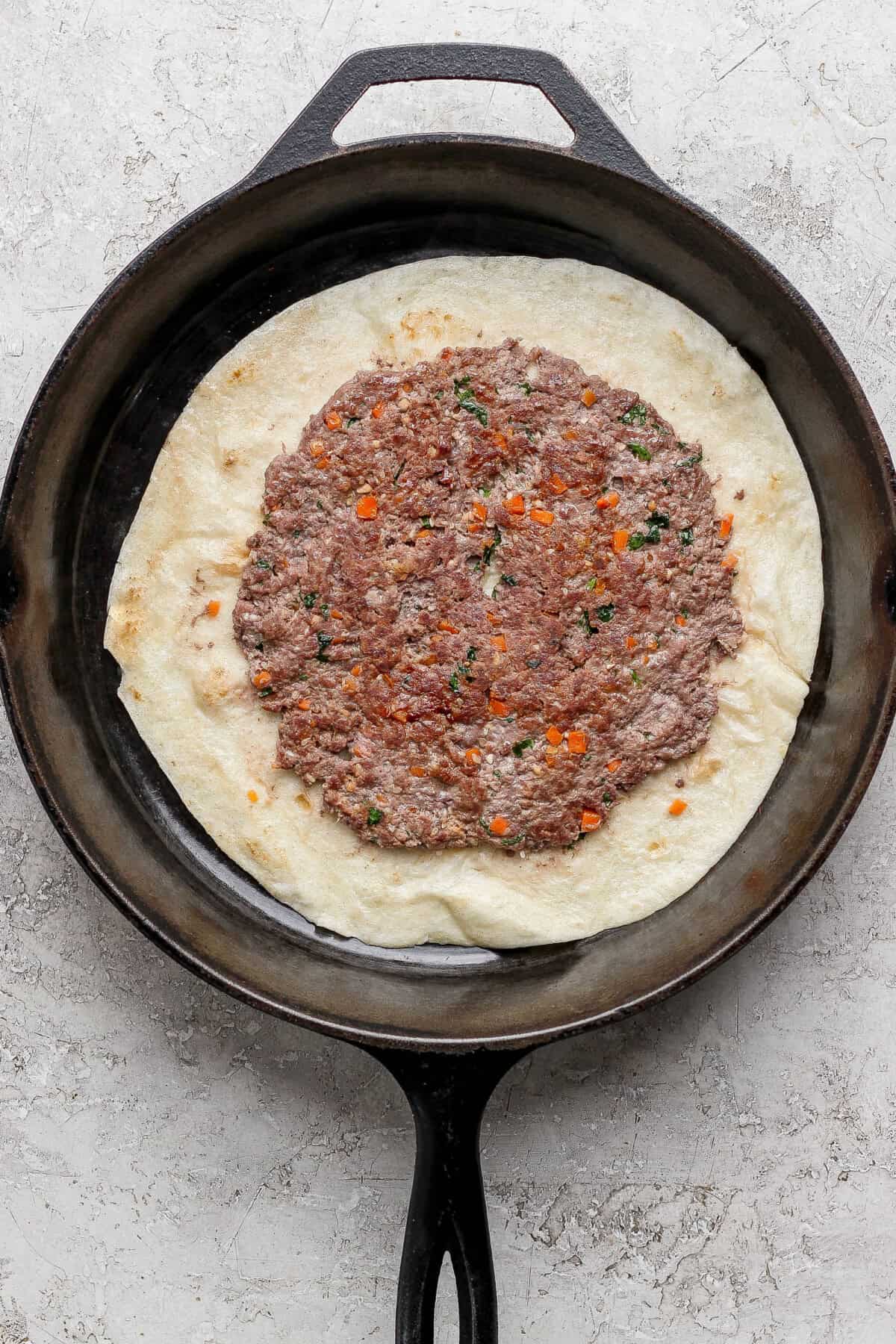 A cast iron skillet with flatbread topped with a spread of seasoned ground meat and chopped vegetables.