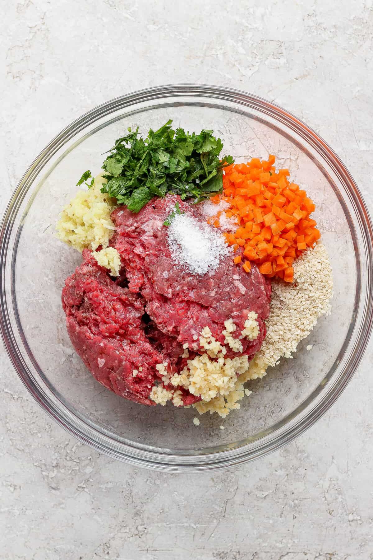 A glass bowl containing ground beef, chopped parsley, diced carrots, minced garlic, breadcrumbs, and salt on a light countertop.